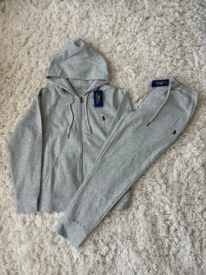 Grått loungewear-set från Polo Ralph Lauren - Mysigt grått loungewear-set från Polo Ralph Lauren med zip-hoodie och matchande byxor. Setet har dragsko i huvan, fickor på tröjan och diskret broderad logga på både tröja och byxa. Perfekt för chill dagar hemma eller när du vill vara bekvämt stylish.