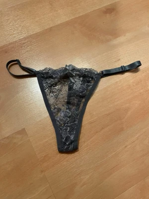 Grå  spetsstring med rosett XL - Säljer en svart stringtrosa i spets med blommigt mönster och liten rosett framtill. Trosan har tunna, justerbara band i sidan och transparent spetsmaterial som ger en sexig look. Perfekt för dig som gillar detaljer och en feminin stil.