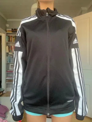 Svart Adidas träningsjacka - Säljer en svart träningsjacka från Adidas med vita ränder längs ärmarna och dragkedja framtill. Jackan har hög krage, logga på båda ärmarna. Perfekt för träning eller chill.