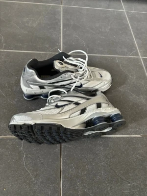 Nike shox silverfärgade sneakers - Snygga sneakers från Nike i silver med svarta detaljer och vit sula. Skorna har en futuristisk design med mesh och syntetmaterial, samt tydliga luftkuddar i hälen för extra komfort. Klassisk Nike Swoosh på sidan och vita skosnören.