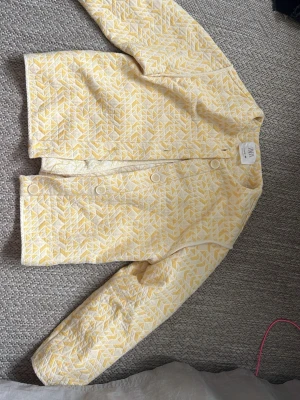 Gulvit quiltad jacka med mönster - Säljer en ljusgul och vit quiltad jacka med geometriskt mönster. Jackan har knappar framtill och lång ärm. Perfekt för dig som vill ha en unik och fräsch look till våren eller hösten.