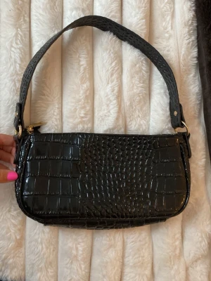Svart axelremsväska i krokopräglat skinn - Snygg svart handväska i krokopräglat konstläder med axelrem och guldfärgade detaljer. Perfekt storlek för det viktigaste och ger en trendig touch till din outfit.🩷🩷köpt för 250kr