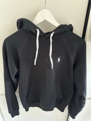 Svart hoodie från Polo Ralph Lauren - Svart hoodie från Polo Ralph Lauren med vit broderad logga på bröstet och vita dragsnören i huvan. Klassisk känguruficka framtill och ribbade muddar vid ärmslut och nederkant. Perfekt för en avslappnad och stilren look. Storlek 160 men fungerar även som xxs