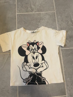 Vit t-shirt med Minnie Mouse-tryck för barn - Vit t-shirt för barn med stort tryck av Minnie Mouse och en katt på framsidan. T-shirten har korta ärmar och rund hals. Trycket är i svart, rosa och lite beige. Perfekt för dig som gillar Disney och vill ha en söt och lekfull stil.