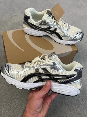 Asics Gel-Kayano 14 Cream/Black - Snygga sneakers från Asics i vitt meshmaterial med silvriga och svarta detaljer. Skorna har en sportig vibe med klassisk Asics-logga på sidan och tjock vit sula. Perfekta för dig som gillar retrostil och bekväma skor med cool design.
