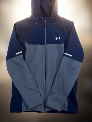 Blå och grå vindjacka från Under Armour - Snygg vindjacka från Under Armour i marinblått och grått. Jackan har hel dragkedja, huva med dragsko och vita detaljer på ärmarna. Tillverkad i ett lätt och vindtätt material, perfekt för blåsiga dagar. Logotypen syns på bröstet.