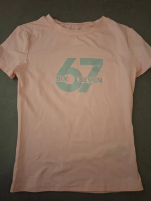 Ljusrosa t-shirt med tryck 67 - Säljer en ljusrosa t-shirt med trycket '67 SIX SEVEN' i vitt framtill. T-shirten har korta ärmar och rund hals. Perfekt för dig som gillar enkel men snygg stil.