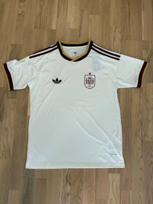  Spanien fotbollströja  - Säljer en vit Spanien fotbollströja från Adidas med vinröda detaljer och guldbruna kanter. Tröjan har korta ärmar, broderad Adidas-logga och Spaniens landslagsemblem på bröstet. Tillverkad i ett lätt och ventilerande material, perfekt för träning eller match.