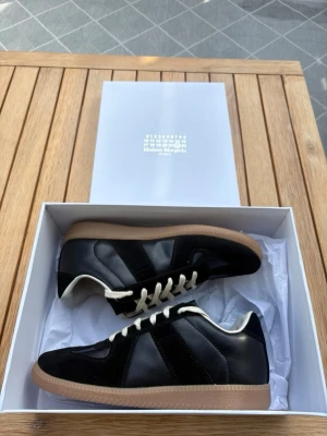 Maison Margiela GAT - Strl 43 - Maison Margiela sneakers i svart läder och mocka med vita snören och beige gummisula. Klassisk low-top siluett med stilrena detaljer och kontrasterande materialval. Perfekta för dig som gillar minimalistisk och trendig design.