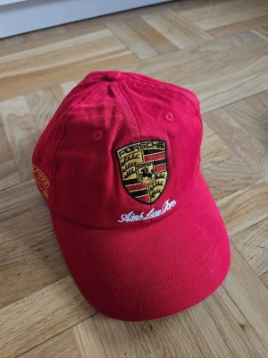 Porsche X Aime Leon Dore Collab Keps - Snygg röd keps med broderad Porsche-logga framtill och vit texten 'Aime Leon Dore' under loggan. Kepsen har böjd skärm och ventilationshål på ovansidan. Tillverkad i bomullstyg och har detaljer i gult och svart.