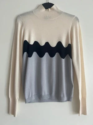 Stickad tröja med vågmönster Marimekko - Snygg stickad tröja med hög krage i beige, ljusgrått och svart. Tröjan har ett vågigt mönster över bröstet och långa ärmar. Materialet är mjukt och tunt. Passar dig som gillar stilrena men unika plagg. Längd ca 60cm. 