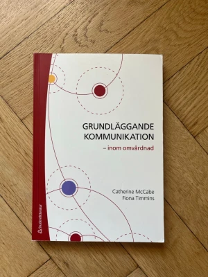 Grundläggande kommunikation inom omvårdnad - Kursbok med vit framsida och röd rygg, titeln 'Grundläggande kommunikation – inom omvårdnad' av Catherine McCabe och Fiona Timmins. Boken är tryckt på Studentlitteratur och riktar sig till studenter inom vård och omvårdnad.