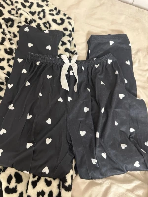 Svarta pyjamasbyxor med hjärtan - Mysiga svarta pyjamasbyxor med vita hjärtan och en dekorativ vit rosett i midjan. Tillverkade i mjukt material med 95% polyester och 5% elastan för extra stretch och komfort. Perfekta för slappa dagar hemma.