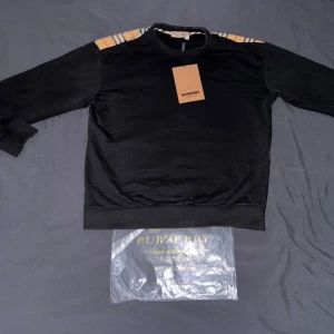 Svart hoodie med rutiga axlar - Snygg svart hoodie från  med klassiska beige, vita och orange rutiga detaljer över axlar och övre rygg. Tröjan har rund halsringning, långa ärmar och ribbade muddar. Perfekt för dig som vill ha en stilren men ändå unik look.