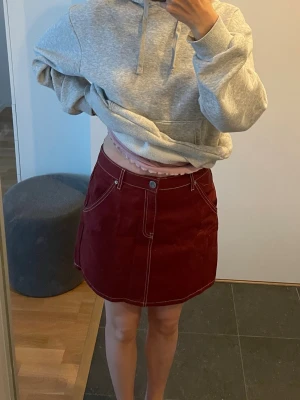 Vinröd jeanskjol med fickor från HM - Vinröd kortkjol från HM i storlek 38 i jeanstyg med vita kontrastsömmar. Kjolen har fram- och bakfickor samt bälteshällor. Aldrig använd. 10 kr!