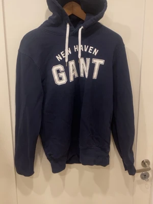 Marinblå hoodie från GANT - Snygg marinblå hoodie från GANT med vit text 'NEW HAVEN GANT' på bröstet. Klassisk huva med vita dragsnören och ribbade muddar vid ärmslut och midja. Perfekt för en avslappnad och sportig stil.