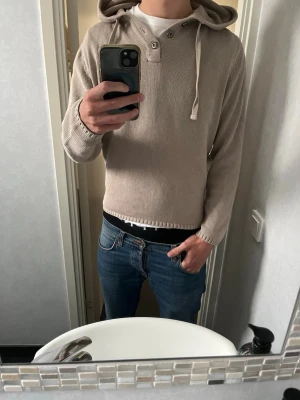 Beige stickad hoodie med huva - Säljer en beige stickad hoodie med huva och dragsko. Tröjan har långa ärmar, ribbade muddar och knappar vid halsen för extra stil. Perfekt för dig som gillar en enkel men snygg look. Passar till jeans eller andra casual outfits.
