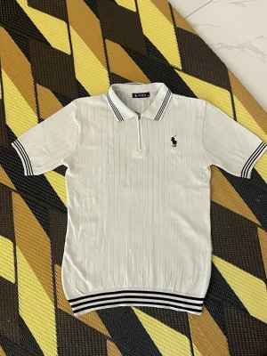 Vit ribbad polotopp från Polo - Vit ribbad polotopp från Polo med svarta ränder på krage och ärmslut. Toppen har korta ärmar, dragkedja framtill och en liten broderad logga på bröstet. Perfekt för en clean och sportig look.