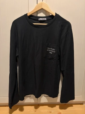 Svart långärmad topp Acne Studios - Svart långärmad topp från Acne Studios med rund halsringning och bröstficka där det står 'Acne Studios STOCKHOLM 1996' broderat i vitt. Enkel och stilren design, perfekt till många olika looks.