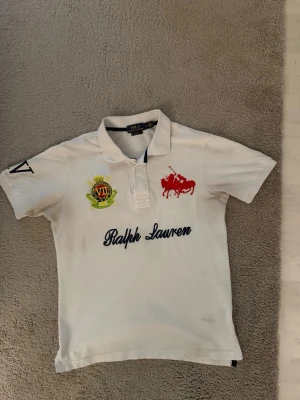 Vit pikétröja från Ralph Lauren - Vit pikétröja från Ralph Lauren med klassisk krage och knappar. Tröjan har broderad logga i rött på bröstet, emblem i grönt och gult samt stor text 'Ralph Lauren' framtill. Kortärmad och i mjuk bomull, perfekt till en avslappnad stil.