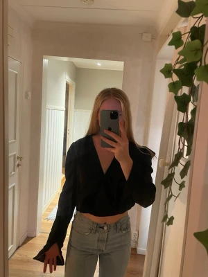 Svart omlottopp med v-ringning - Snygg svart omlottopp med v-ringning och långa ärmar. Toppen är croppad och har en stilren look som passar perfekt till jeans eller kjol. Materialet känns mjukt och lätt, vilket gör den bekväm att bära.
