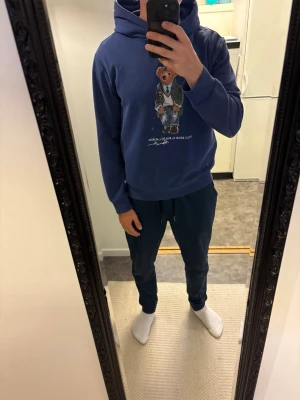 Blå hoodie med Polo Bear från Ralph Lauren - Snygg blå hoodie från Ralph Lauren med den ikoniska Polo Bear-trycket på bröstet. Hoodien har huva med dragsko och ribbade muddar vid ärmar och nederkant. Modellen på bilden är 182 och väger 80kg. Köpt ny för 2395kr, endast använd fåtal gånger.