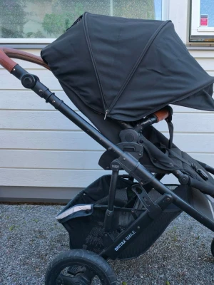 Svart barnvagn Britax Smile - Säljer en svart barnvagn Britax Smile med robust ram i metall och detaljer i brunt konstläder på handtaget. Vagnen har stora hjul, rymlig varukorg undertill och justerbart ryggstöd. Perfekt för promenader och enkel att manövrera.