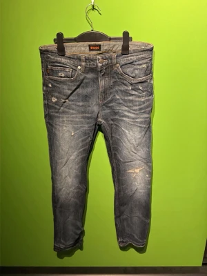 Blå slitna jeans från BOSS - Säljer ett par blå jeans från BOSS med slitningar och distressed detaljer. Klassisk femficksmodell med normal midja och raka ben. Jeansen har en cool tvättad look och orange sömmar som kontrast. Perfekta för en avslappnad streetstyle.
