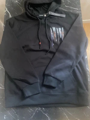 Hoodie AMIRI MA Bar Logo Drip Noir M 🎨 - Svart hoodie från Amiri med huva och dragsko. På bröstet finns ett vitt Amiri-tryck med färgstänk i blått och orange. Tillverkad i mjukt material och har ribbade muddar vid ärmar och nederkant. Perfekt för en streetwear-look.