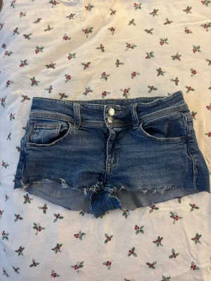 Blå denimshorts med rå kant - Snygga blå denimshorts med låg midja och rå, fransig kant. Klassisk femficksmodell med dubbla knappar framtill och fickor med lock bak. Perfekta för varma dagar och enkel att matcha med allt. Orginellt jeans från ginatricot som man sedan klippt till shorts, Jeans storlek 164.