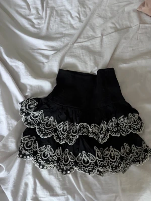 Svart volangkjol med spetsdetaljer - Supersöt svart kortkjol med flera lager volanger och vita broderade spetsdetaljer. Kjolen har hög midja och ger en lekfull och trendig look, perfekt att styla med en enkel topp.