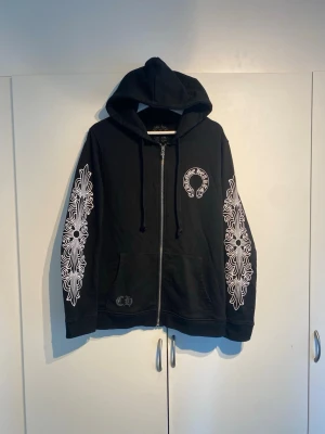Chrome Hearts Zip Hoodie  - Säljer en svart Chrome Hearts zip hoodie med dragkedja och huva. Tröjan har vita grafiska mönster längs båda ärmarna och en rund logga på bröstet. Fickor framtill och snörning i huvan. Perfekt för dig som gillar streetwear och snygga detaljer. Inte 100% på äkthet, därav priset. Storlek M med avslappnad passform. Sparsamt använd, fint skick.