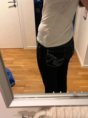 Svarta jeans med vita detaljer - Säljer ett par svarta jeans med snygga vita sömmar och dekorativa nitar på bakfickorna. Jeansen har en klassisk femficksmodell och normal midja. Passar perfekt till dig som vill ha en stilren men ändå unik look.