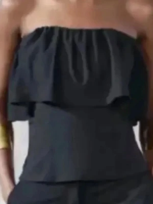 Svart axelbandslös volangtopp - Snygg svart axelbandslös topp med volangdetalj upptill. Perfekt för dig som gillar en stilren och trendig look. Toppen är ärmlös och har en bandeauform som sitter snyggt över axlarna.helt slutsåld.