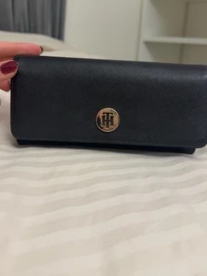Svart plånbok från Tommy Hilfiger - Snygg svart plånbok från Tommy Hilfiger i ett tåligt material med guldfärgad logga framtill. Plånboken har flera kortfack, myntfack med dragkedja och tryckknappsstängning. Perfekt för att hålla ordning på kort  och kontanter.