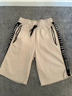Beige shorts med svarta detaljer - Snygga beige shorts med svarta snören i midjan och svarta sidopaneler med vit text. Shortsen har två dragkedjeförsedda fickor framtill och elastisk midja för skön passform. Perfekta för en avslappnad stil.