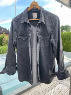 Mörkgrå overshirt i denim - Säljer en snygg mörkgrå overshirt i denim-look med klassisk krage och två bröstfickor med knappar. Skjortan har långa ärmar och knäppning framtill. Perfekt för dig som gillar en avslappnad och trendig stil.