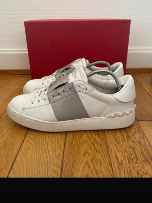 INTRESSEKOLL!!!!! Valentino garvani skor - Snygga vita sneakers från Valentino med grått band över mitten och coola nitar på hälen. Skorna har en klassisk låg profil, vita snören och detaljer i läder. Perfekta för dig som vill ha en stilren men ändå unik look. Box följer inte med! Skorna har även ett hål på vänster sko - se sista bild! Man ser inte hålet när man har på sig dom!