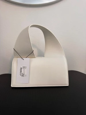 Vit minimalistisk handväska - Stilren vit handväska i slät konstläder med unik, böjd form och bred axelrem. Väskan har en modern och minimalistisk design utan synliga detaljer eller loggor. Perfekt för dig som gillar trendiga accessoarer med enkel look.