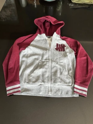 Grå och vinröd hoodie från Undefeated - Säljer en grå hoodie med vinröda ärmar och huva från Undefeated. Dragkedja framtill, två fickor och ikoniskt logotryck på bröstet. Ärmsluten har vinröda och vita ränder. Perfekt för en avslappnad streetwear-look.