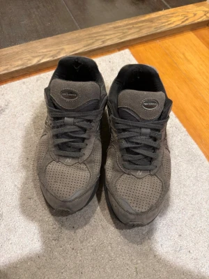 Grå New Balance sneakers i mocka - Säljer ett par grå New Balance sneakers i mocka med svarta detaljer och perforerat mönster på tån. Klassisk låg modell med svart sula och snörning. Perfekta för dig som gillar stilrena och bekväma skor. The back of the shoe is broken inside, which is included in the price. 