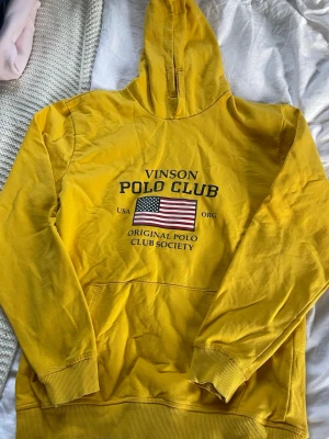 Gul hoodie från Vinson Polo Club - Snygg gul hoodie från Vinson Polo Club med stor amerikansk flagga och text på bröstet. Klassisk känguruficka framtill och ribbade muddar vid ärmar och nederkant. Tillverkad i mjukt bomullsmaterial och har en stor huva för extra chill vibe.