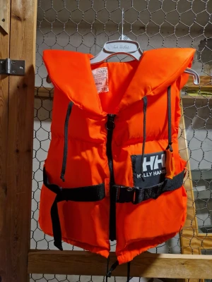 Orange flytväst från Helly Hansen - Säljer en klassisk orange flytväst från Helly Hansen med svart bälte och dragkedja framtill. Västen har en stor krage och tydlig logga på bröstet. Perfekt för alla vattenaktiviteter där säkerhet är viktigt. Vikt 40-60kg. Använd 1 gång.