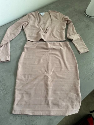 Beige set från NLY One med kjol och topp - Säljer ett beige tvådelat set från NLY One i storlek S. Setet består av en långärmad topp med djup v-ringning och en matchande midilång kjol. Materialet är mjukt och stretchigt med diskreta horisontella ränder. Perfekt för dig som gillar stilrena och trendiga plagg.