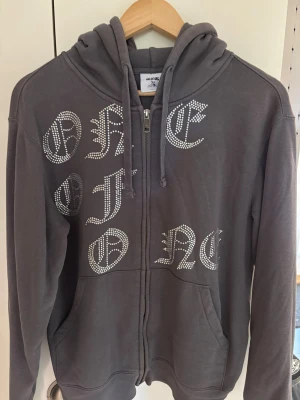 Grå hoodie med dragkedja och tryck - Snygg grå hoodie från One of One med dragkedja och huva. Framsidan har ett coolt tryck i stora bokstäver med glittrande stenar. Hoodien har fickor framtill och justerbara snören i huvan. Perfekt för en avslappnad streetwear-look.