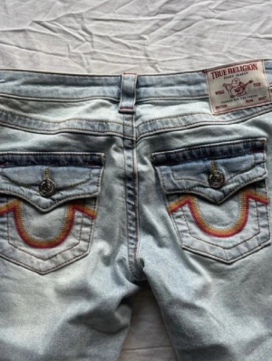 True Religion ljusblå jeans med brodyr - Säljer ett par ljusblå jeans från True Religion med ikoniska broderade fickor i rött och gult. Jeansen har raka ben och klassisk passform. Snygga detaljer med knappar på bakfickorna och tydlig logopatch i midjan.