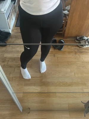 Svarta Nike leggings med logga - Säljer ett par svarta leggings från Nike med klassisk vit logga på ena benet. De har en tight passform och hög midja, perfekta för träning eller chill. Materialet är stretchigt och följsamt för maximal komfort.