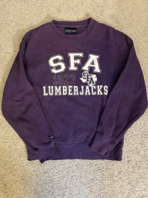 Lila SFA Lumberjacks sweatshirt - Lila sweatshirt från Jansport med tryck 'SFA 1923 Lumberjacks' i vitt och lila på bröstet. Rund halsringning och långa ärmar. Köpt på en finare vintage second hand butik i Stockholm. 