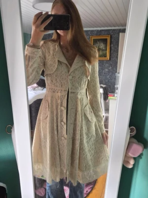 Krämvit spetskappa med volangkrage - Säljer en elegant krämvit kappa i spets med volangkrage och dekorativa knappar framtill. Kappan har markerad midja, fickor och ett utsvängt fall som ger en romantisk look. Perfekt för dig som vill sticka ut med en unik och feminin stil.
