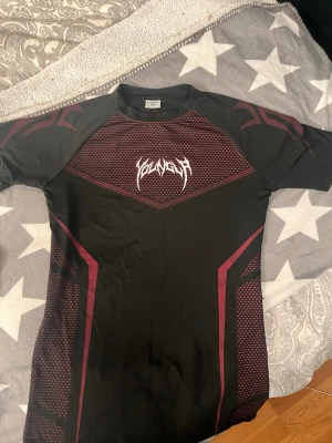 Compression shirt YoungLA - Säljer en svart compression shirt från YoungLA i storlek S. Tröjan har röda grafiska detaljer och ett vitt tryck på bröstet. Tillverkad i ett stretchigt och snabbtorkande material, perfekt för träning eller gym. Snygg passform och korta ärmar. Org påsen följer med